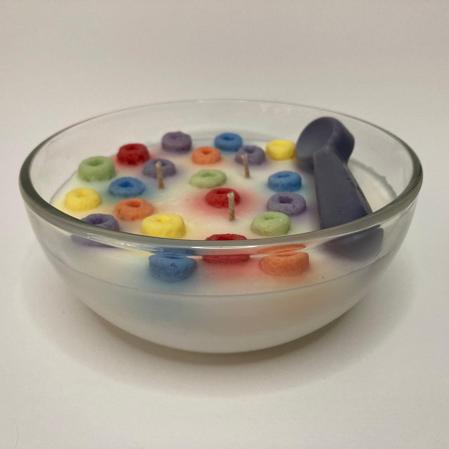 A glass bowl filled with a handcrafted Loopty Fruity scented candle designed to look like a bowl of milk and colorful ring-shaped cereal, complete with small wicks and a silver spoon.
