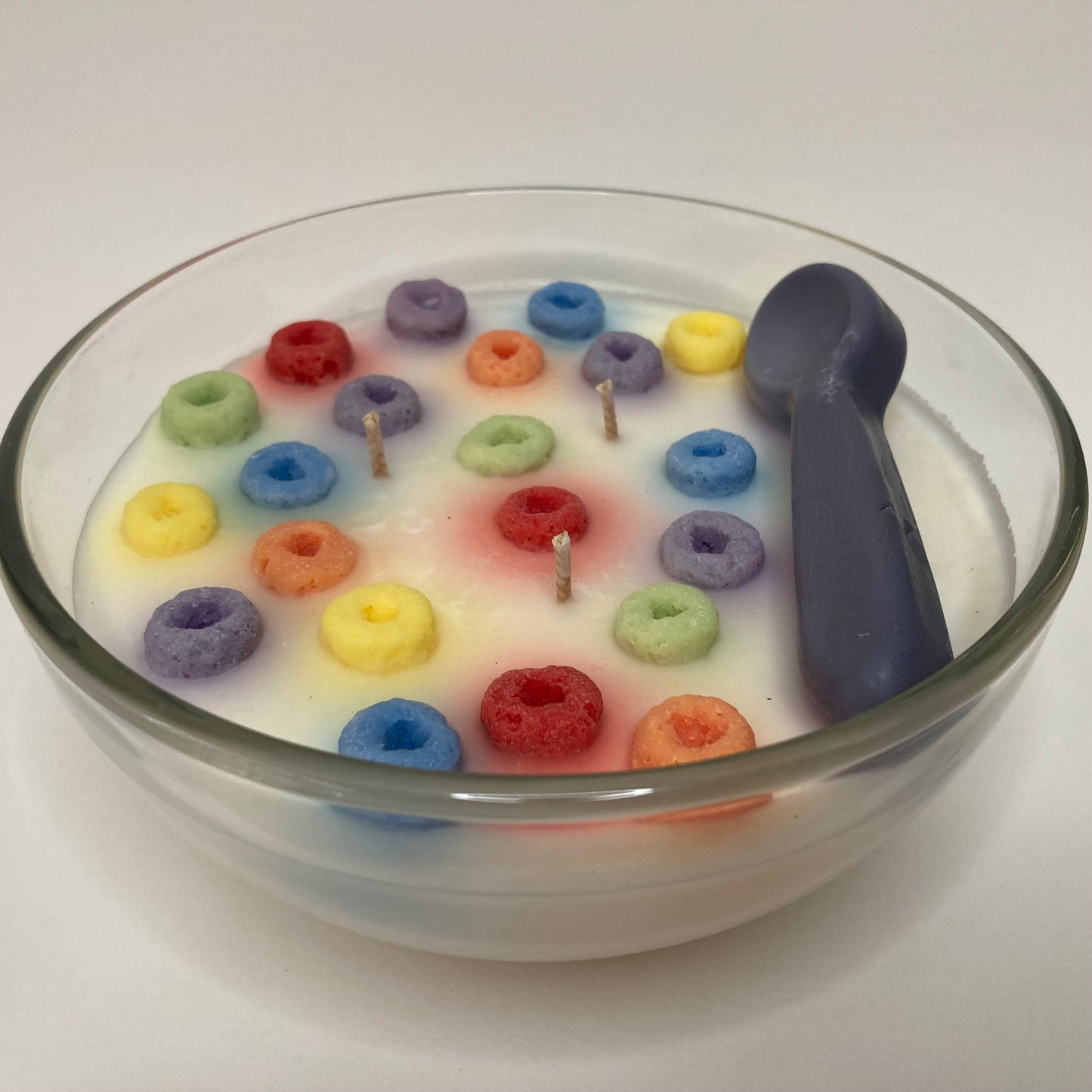 Loopty Fruity cereal bowl candle. A realistic clear glass bowl filled with colorful ring cereal pieces in milk-white scented wax, complete with three wicks and a silver spoon.
