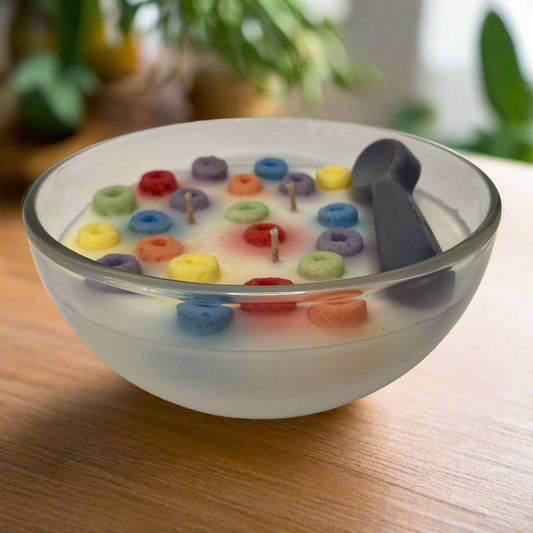 Detailed view of the A Happy Waxident 'Fruit Loop' scented cereal bowl candle, featuring multi-colored fruit-loops in a glass dish with wicks and a spoon, resting on a wood table.