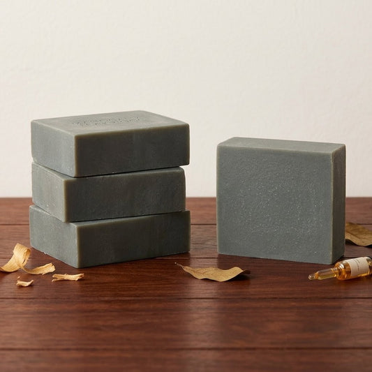A neat stack of three handcrafted, green Mahogany Teakwood donkey milk soap bars next to a single upright bar, accompanied by wood shavings, a dried leaf, and a small glass vial on a warm wooden table.