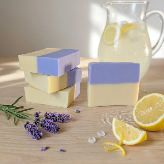 A rustic stack of handcrafted Lavender Lemonade donkey milk soap bars next to a glass pitcher of iced lemonade and freshly cut lemon slices.