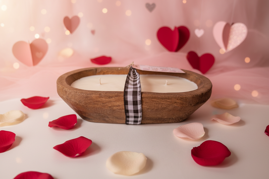 A Happy Waxident "Love Spell" dough bowl candle. Rustic hand-carved wooden bowl filled with multi-wick soy wax and wrapped with a black and white gingham ribbon. The romantic scene includes scattered rose petals and heart decor.