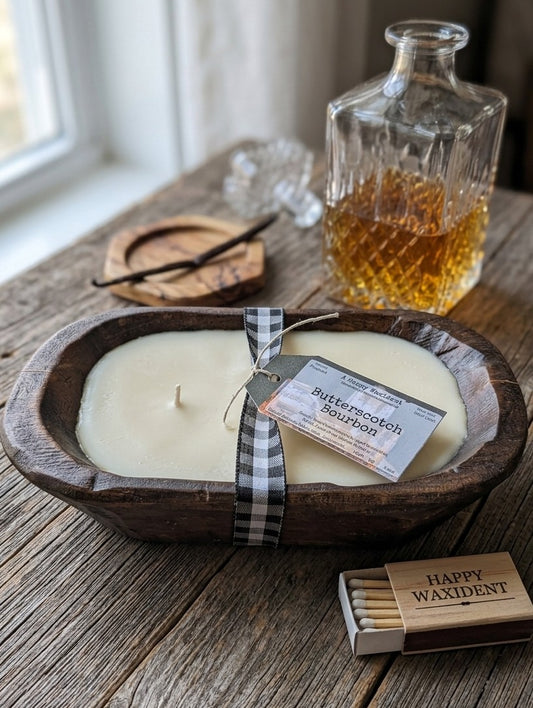 Rustic, dark wood 2-wick dough bowl candle with Butterscotch Bourbon fragrance by A Happy Waxident. The candle is wrapped in a black and white gingham ribbon with a detailed product tag. It is presented on a weathered wood table with complementary lifestyle props including a crystal decanter with amber bourbon, a whole vanilla bean on a wood coaster, and a branded matchbox, showcasing the farmhouse aesthetic and warm scent notes